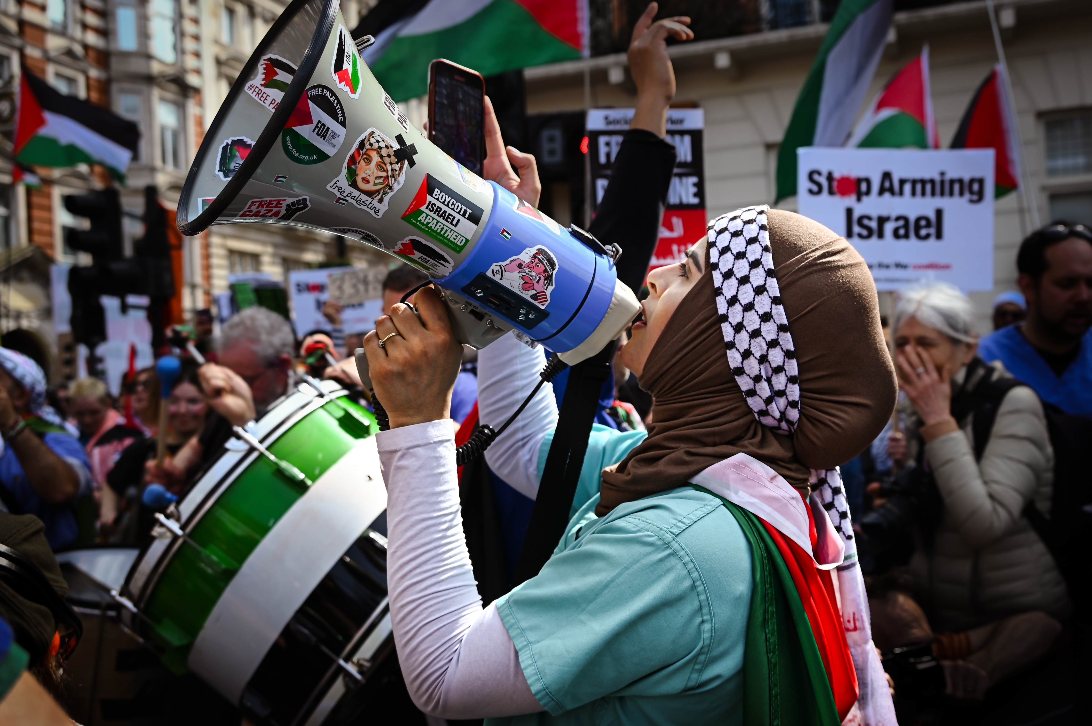 A woman stands shouting into a megaphone. She is draped in a Palestinian flag, and behind her are others holding up banner saying Stop Arming Isreal with more Palestinian flags. 