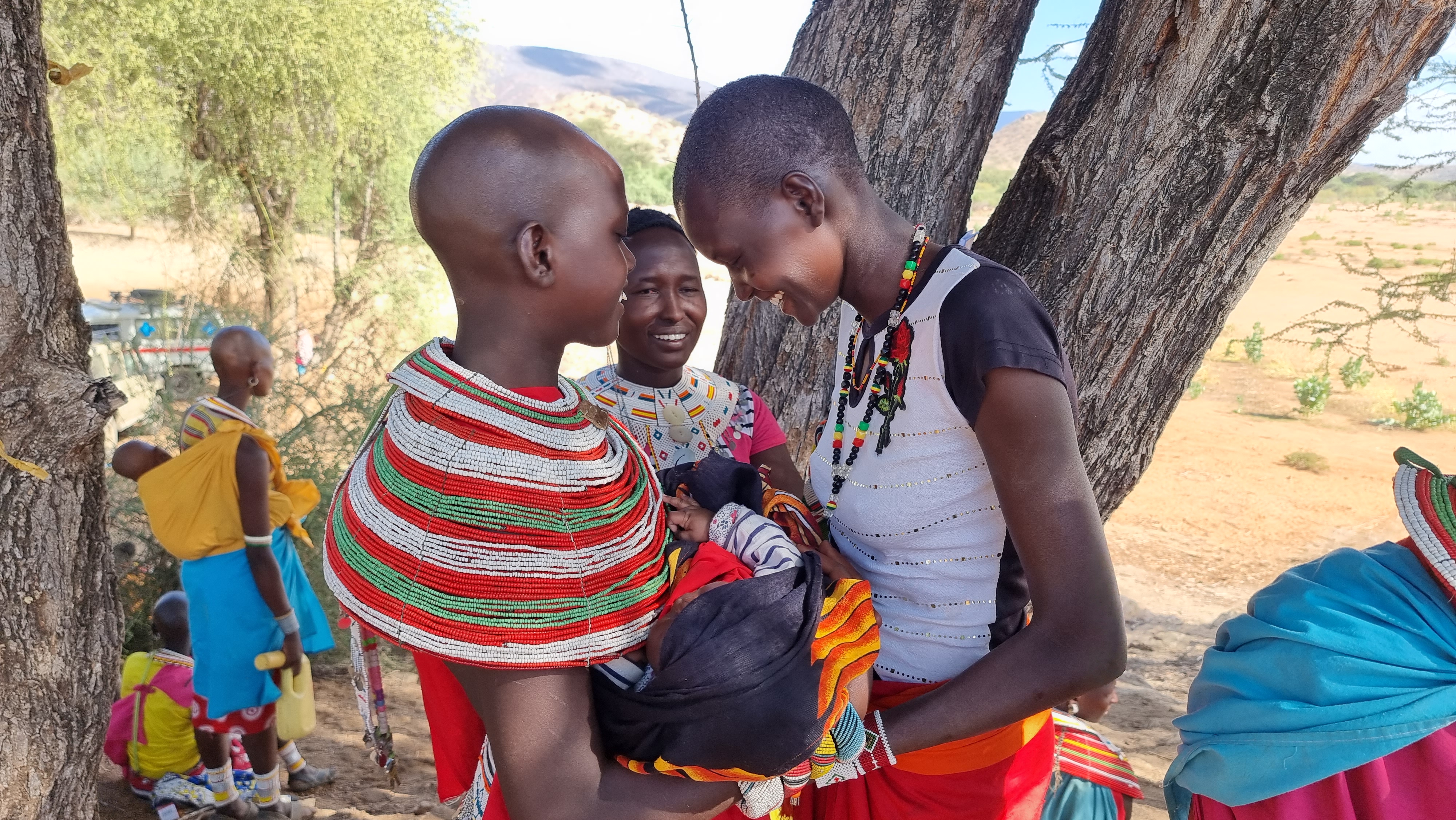Three women stand over a baby in traditional dress in Kenya. They stand with their heads close together smiling. 