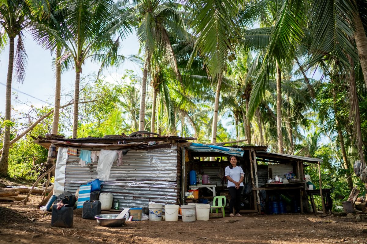 A woman stands in front of her makeshift home surrounded by palm trees