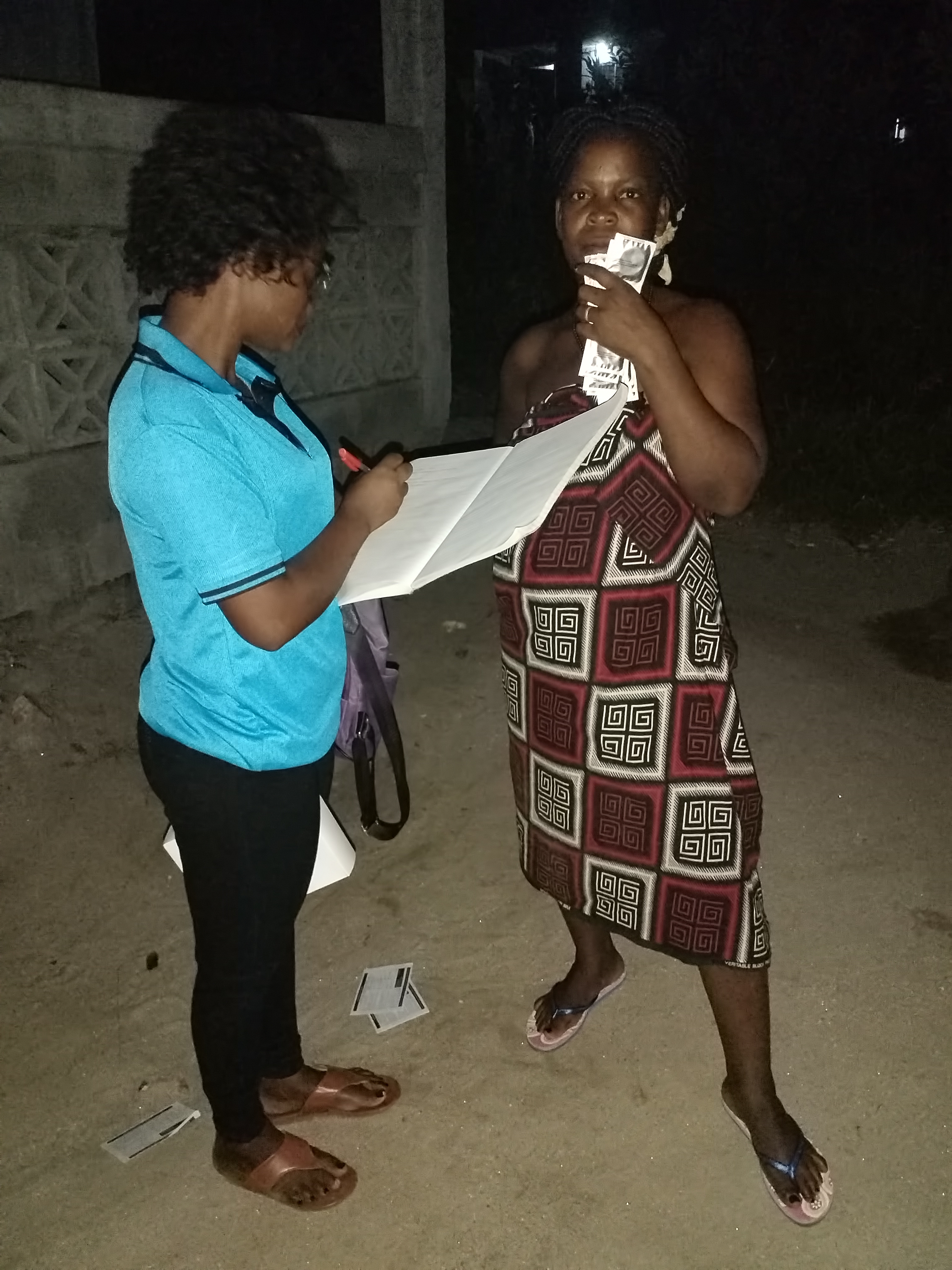 Two women stand in front of the camera. One of the women who a peer educator is filling out a form. The other woman is a sex worker and is holding a pack of condoms in her hand. 