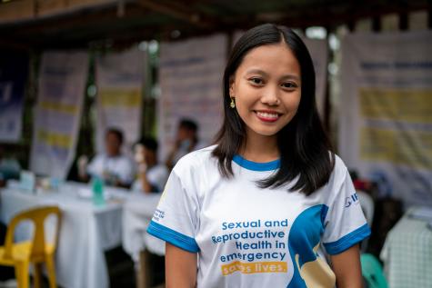 A women stands facing the camera smiling wearing a t-shirt that reads "Sexual and Reproductive Health in Emergency Saves Lives"