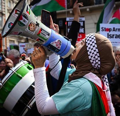 A woman stands shouting into a megaphone. She is draped in a Palestinian flag, and behind her are others holding up banner saying Stop Arming Isreal with more Palestinian flags. 
