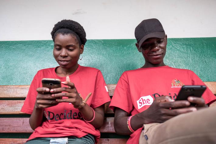 The image shows two people in red t-shirts both holding a mobile phone.