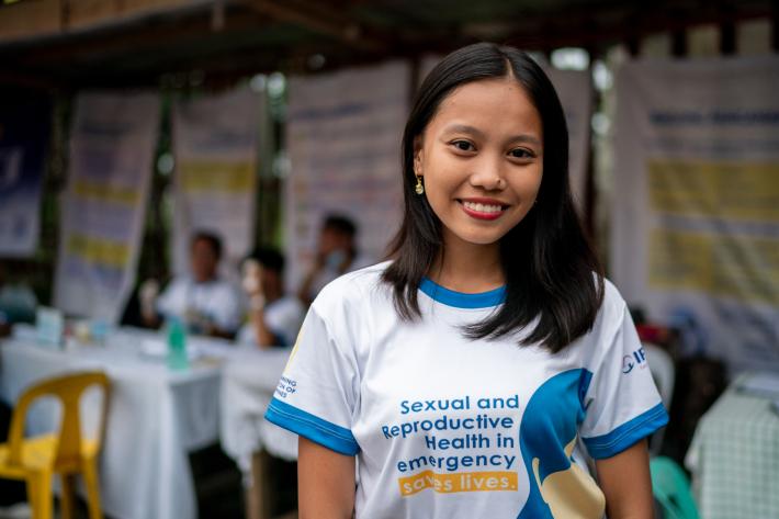 A women stands facing the camera smiling wearing a t-shirt that reads "Sexual and Reproductive Health in Emergency Saves Lives"