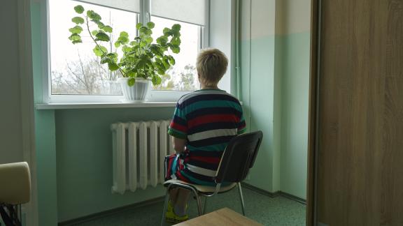 A Ukrainian women sits in a chair with her back to the camera facing the window. There is a green plant on the window ledge, and the room is painted a light green. 