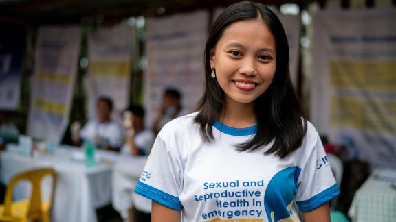 A women stands facing the camera smiling wearing a t-shirt that reads "Sexual and Reproductive Health in Emergency Saves Lives"